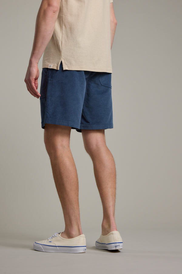 A person stands facing away, arms at their sides, against a plain light background wearing a beige short-sleeve shirt, Barkers Montauk Cord Weekend Short with an elastic waistband, and beige canvas sneakers.