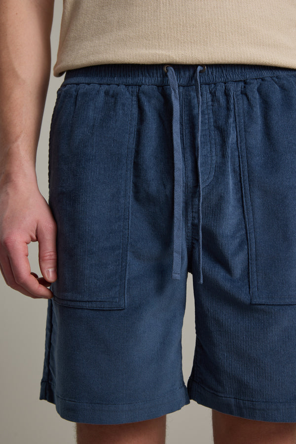 A person wears blue Barkers Montauk Cord Weekend Shorts featuring an elastic waistband and drawstring. The image, cropped to the lower torso and upper legs, shows one arm relaxed by their side.
