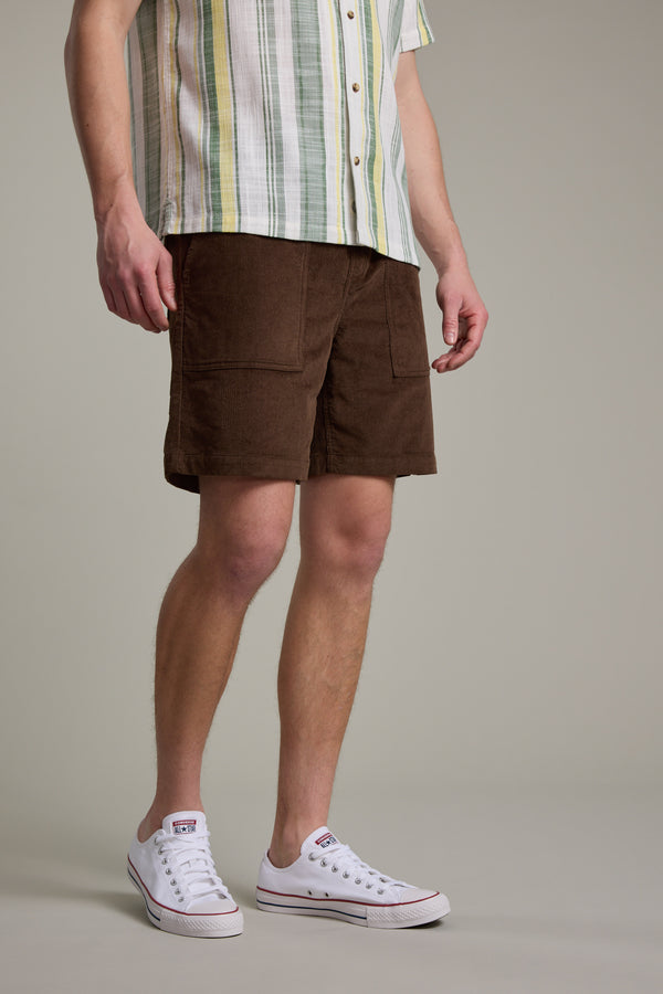 A person wears a short-sleeve, striped button-up, Barkers Montauk Cord Weekend Short with an elastic waistband, and white low-top Converse sneakers. The chest-cropped photo features a neutral background.