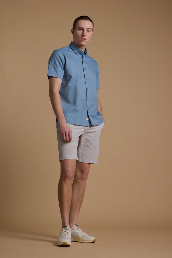 A young man stands against a beige backdrop, wearing the Barkers Archer Oxford Shirt in light blue, paired with beige shorts and light sneakers. With one hand in his pocket, he gazes slightly to the side.