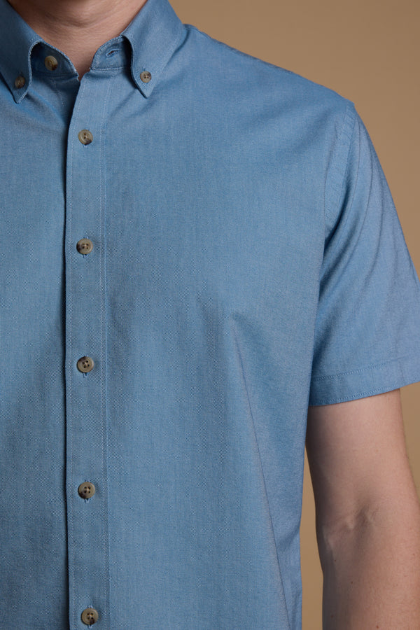 A person wears the Barkers Archer Oxford Shirt, a light blue, short-sleeve, 100% cotton top with a button-down collar, shown from shoulders to mid-torso against a plain beige background.