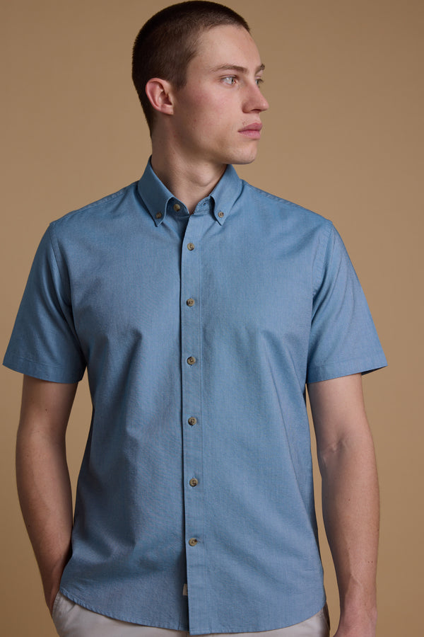 A man stands against a beige background wearing the Barkers Archer Oxford Shirt in light blue, featuring short sleeves and a button-down collar, while looking to his left with a neutral expression.