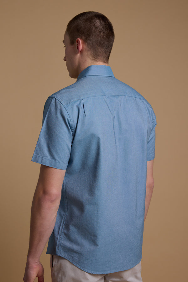 A man stands with his back to the camera against a plain beige background, wearing the Barkers Archer Oxford Shirt in light blue with short sleeves and a button-down collar, paired with light-colored pants.