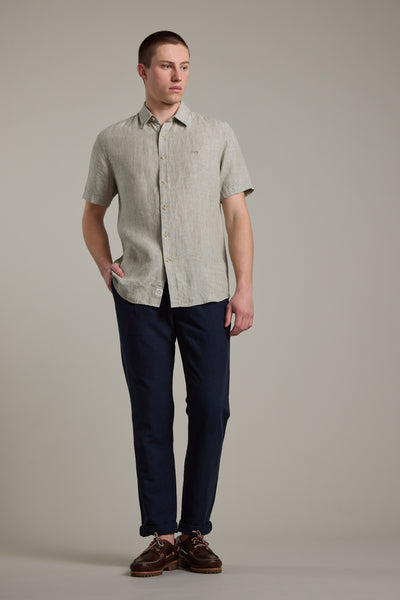 A man stands against a plain background wearing Barkers’ Newman Linen Pant in dark navy with a light beige short-sleeve button-up shirt and brown shoes. He has one hand in his pocket and gazes slightly to the side.