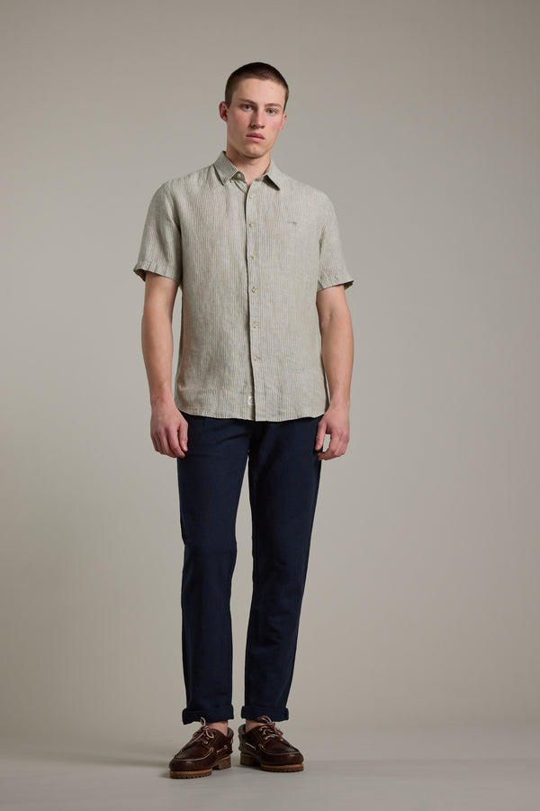 A young man stands against a light grey background, wearing Barkers Newman Linen Pant in dark blue with a short-sleeved light beige shirt and brown shoes. He faces forward, maintaining a neutral expression.