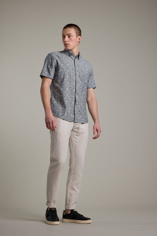 A man stands before a plain background in the Barkers Shelby Print Shirt—a short sleeve summer shirt—paired with light beige pants and black sneakers, looking to the side with a neutral expression.