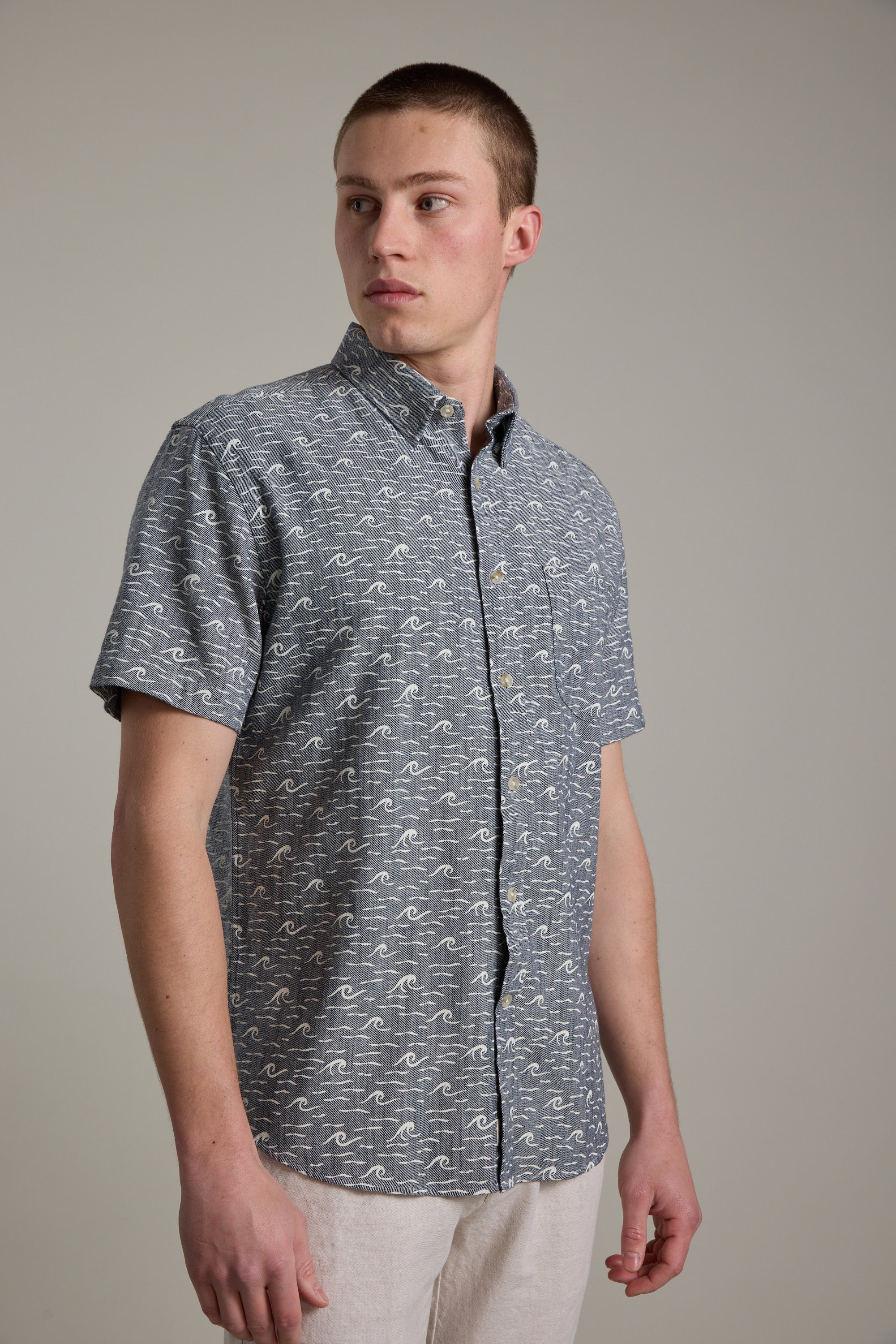 A young man with short hair models the Barkers Shelby Print Shirt, a gray short sleeve summer shirt with a white wave pattern, made from responsibly sourced cotton. He stands against a plain light gray background, looking slightly to his left.