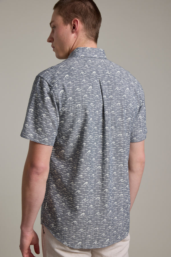 A man with short brown hair faces slightly left, back to the camera, wearing a Barkers Shelby Print Shirt—a short sleeve gray shirt with a white wave pattern, made from responsibly sourced cotton—against a plain background.