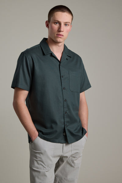 A young man with short hair stands against a plain background, wearing the Barkers Augusta Cuban Shirt in dark green—a boxy-fit style made from responsibly sourced cotton—and light gray pants, with his hands in his pockets.