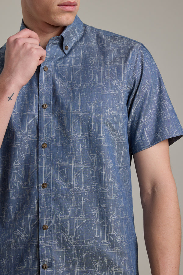 A man models the Barkers Yacht Jacquard Shirt, a blue short sleeve cotton shirt with a subtle yacht print. His left hand, showing a small cross tattoo on his wrist, is near his chin. The background is plain and neutral.