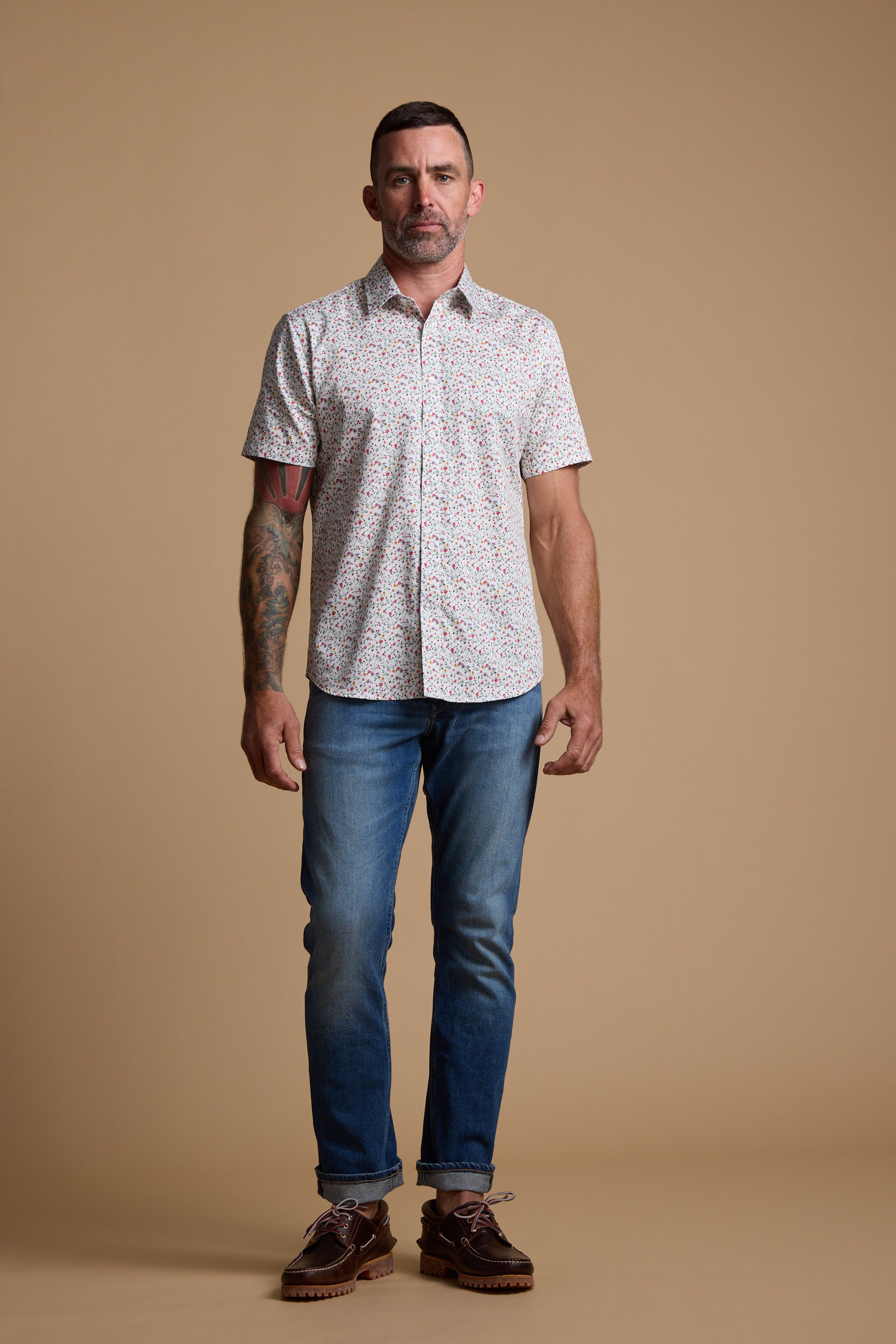 A man with short dark hair, a trimmed beard, and a left forearm tattoo stands against a beige background wearing the Barkers Wade Print Shirt—short-sleeve floral print made from responsibly sourced cotton—paired with blue jeans and brown shoes.