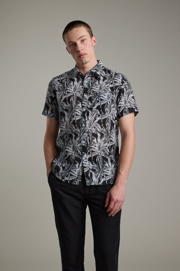 A young man stands against a plain background, wearing the Barkers Madison Print Shirt—a short-sleeved, black cotton–linen blend with a white tropical leaf pattern—and black pants. He has short hair, a relaxed expression, and one hand in his pocket.