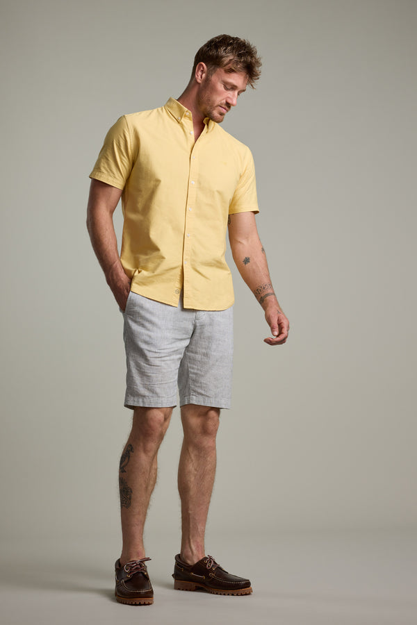 A man stands against a plain background wearing Barkers’ Palma Walkshorts in light gray, a yellow button-up shirt, and brown boat shoes. With one hand in his pocket, he looks down to his left, showcasing relaxed summer style.