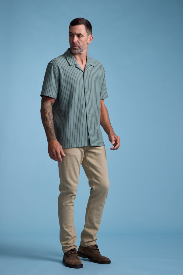 A man with short dark hair and tattoos on his left arm stands against a light blue background, wearing the Barkers Diego Stripe Cuban Shirt, beige pants, and brown shoes.