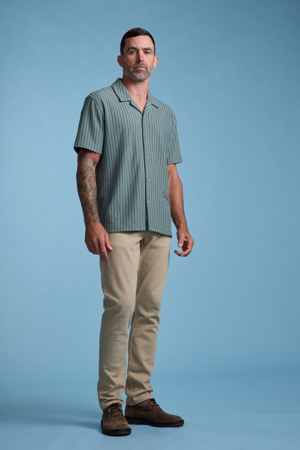 A man with short dark hair and tattoos on his right arm stands against a blue background, wearing the Barkers Diego Stripe Cuban Shirt, which has a boxy fit, paired with beige pants and brown shoes.
