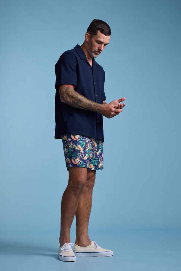 A man stands against a light blue background, wearing the Barkers Diego Cuban Shirt, colorful tropical-patterned shorts, and white sneakers. He looks down, appearing to touch his hands together.