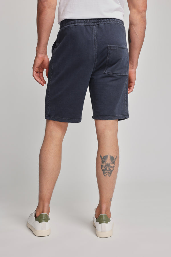 A person shown from the back wears a white shirt, Barkers Organic Classic Print Track Short in navy blue, and white sneakers with green accents. A stylized face tattoo appears on their left calf. The shorts display the Barkers heritage logo.