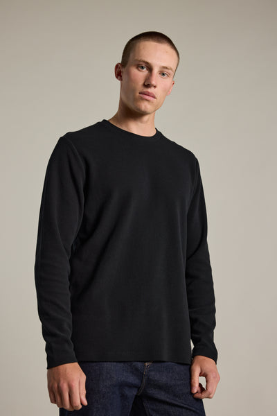 A young man with short hair stands against a neutral background, wearing the Barkers Avenue Waffle Crew long-sleeve shirt in black and dark jeans, looking directly at the camera with a neutral expression.
