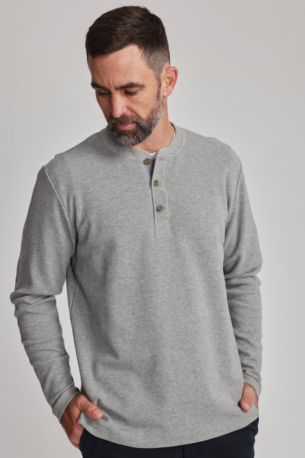 A man with short dark hair and a beard wears the Barkers Avenue Waffle Henley, a mid-weight organic cotton shirt with a subtle waffle texture. He looks down, hands in his pockets, against a plain light background.