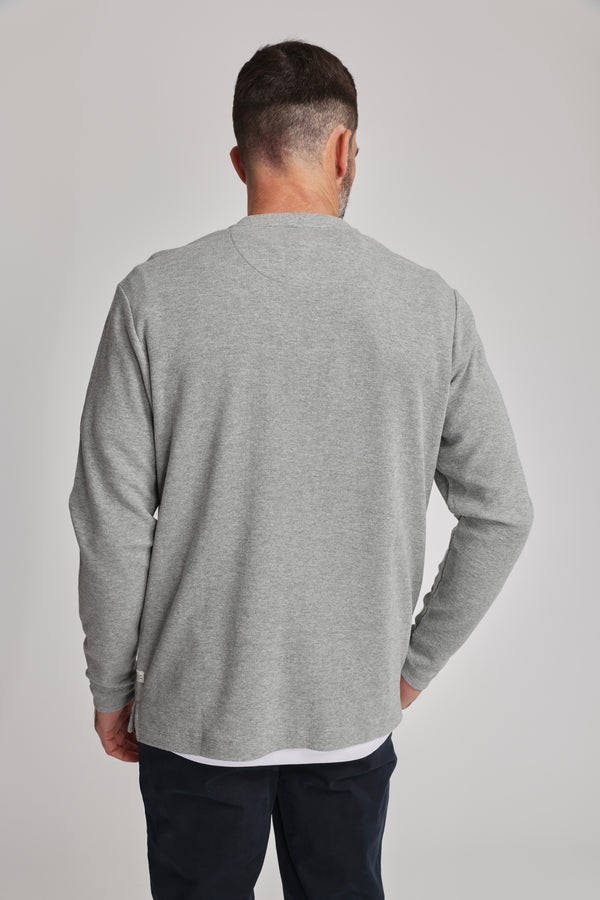 A man with short dark hair and a trimmed beard stands with his back to the camera, wearing the Barkers Avenue Waffle Henley in light gray and dark pants against a plain light background.