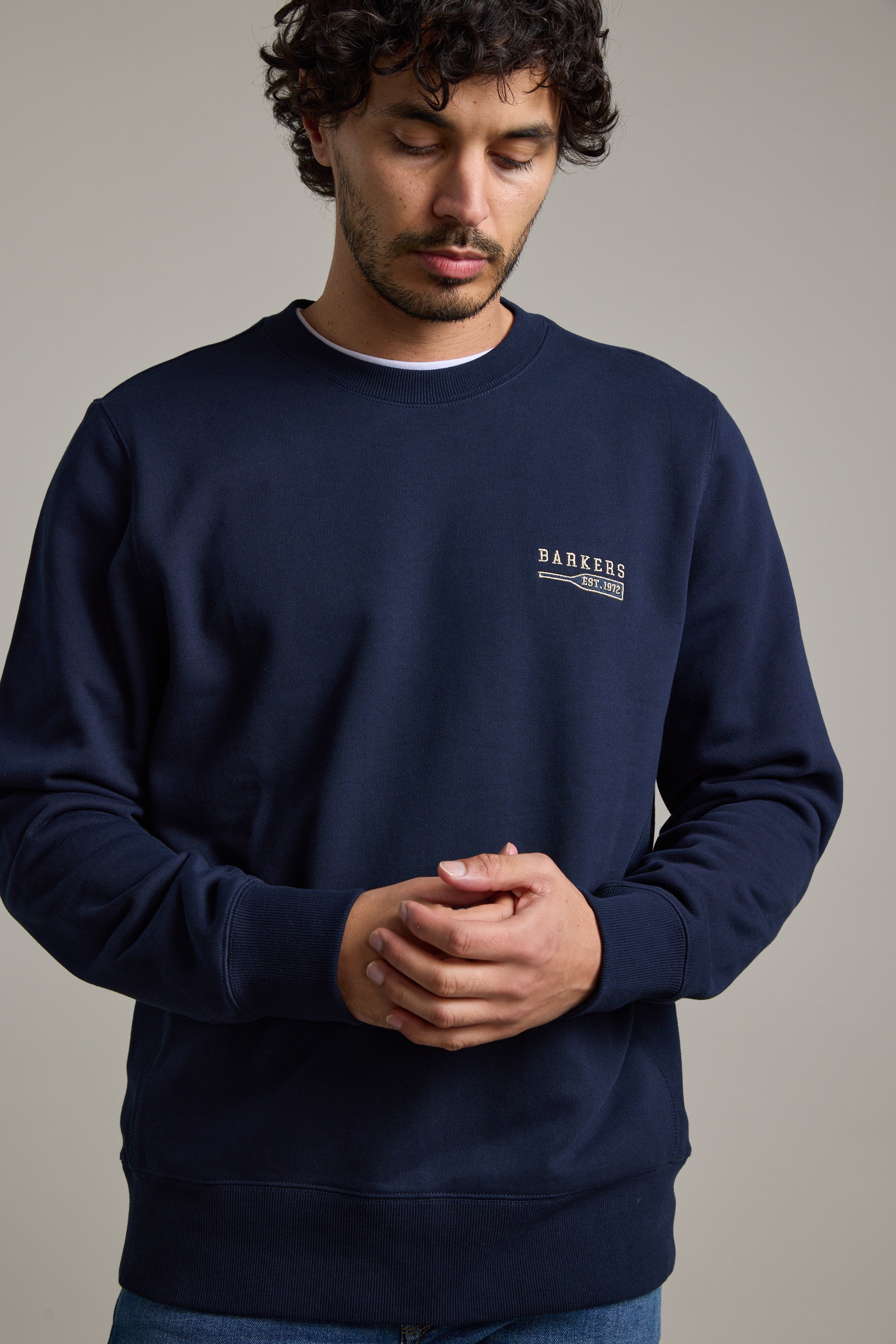 A man with curly hair wears the Barkers Regatta Crew sweatshirt by Barkers, made from responsibly sourced cotton, standing against a plain background and looking down with his hands clasped.