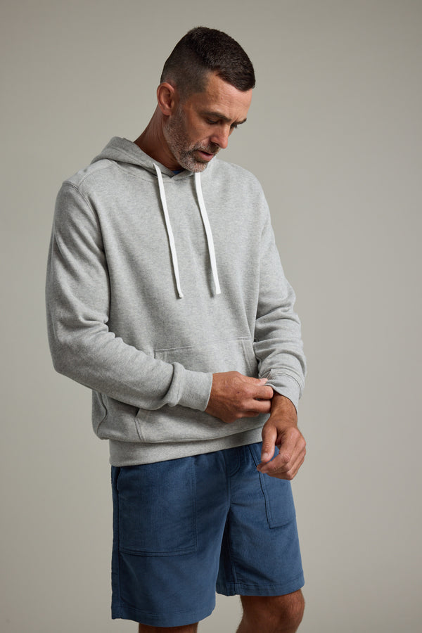 A man wearing the Barkers Inlet Hoody in light gray and blue shorts looks down, adjusting his sleeve against a plain, light background.