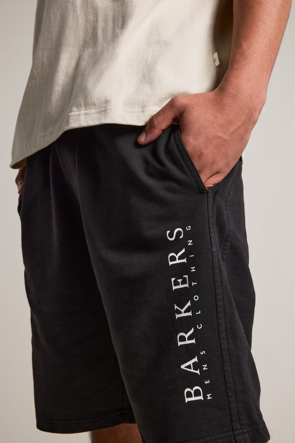 Someone wears Barkers Organic Classic Print Track Shorts in black, featuring the heritage logo on the side, paired with a light t-shirt. One hand is in their pocket against a plain, neutral background.