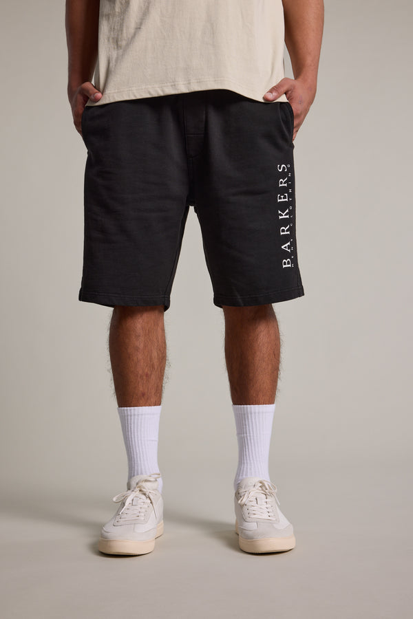 A person wearing a beige T-shirt, Barkers Organic Classic Print Track Short in black with white lettering on the left leg, white crew socks, and white sneakers stands against a plain light gray background.