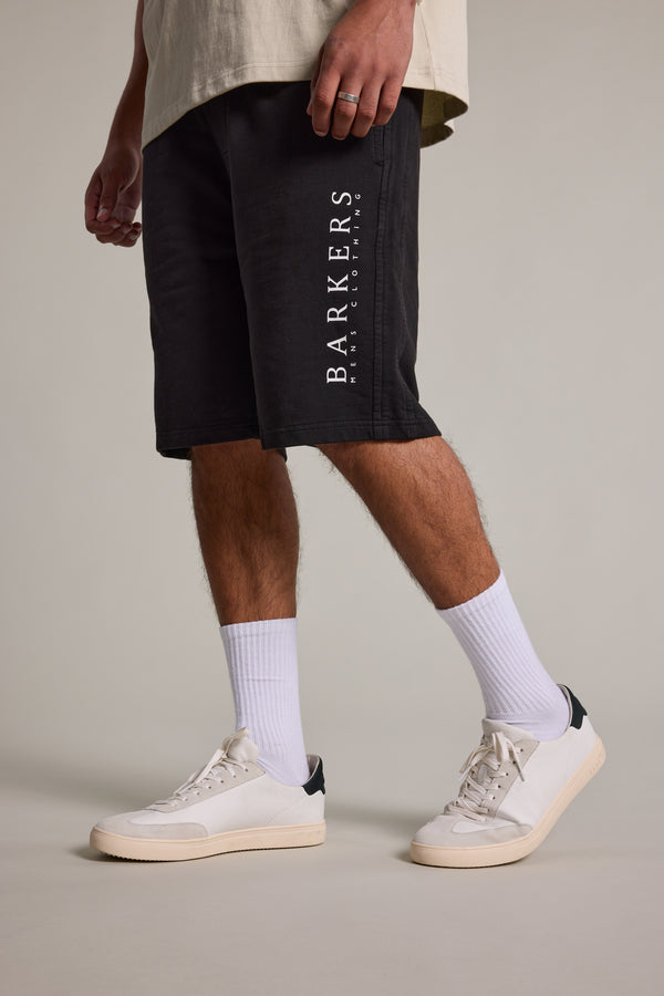 A person stands against a plain background, wearing Barkers Organic Classic Print Track Shorts in black, paired with white crew socks and white sneakers. Only the lower half of their body is visible.
