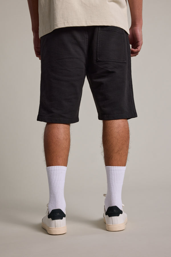 A person stands facing away, wearing Barkers Organic Classic Print Track Shorts in black, a beige T-shirt, white crew socks, and white sneakers with dark green heels, set against a plain light background.