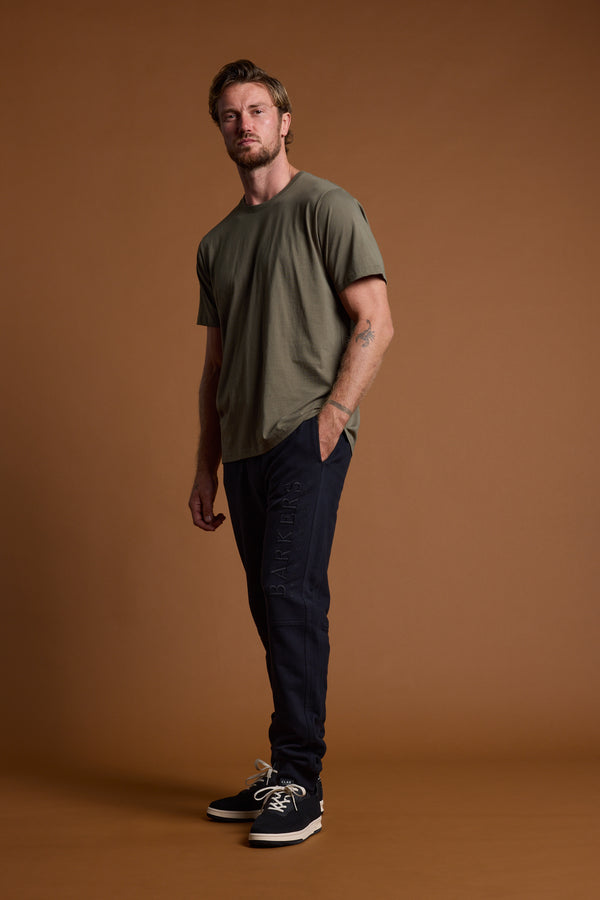A man with short hair, a beard, and a tattoo on his left forearm stands against a brown background wearing an olive green t-shirt, Barkers Heritage Track Pant by Barkers, and black sneakers.