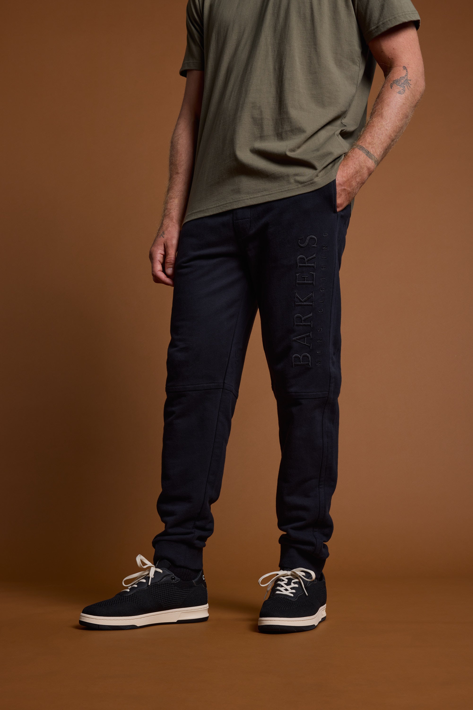 A man, face unseen, stands against a brown background wearing a green tee and Barkers Heritage Track Pant with BARKERS branding on the leg, paired with black sneakers with white soles and laces.