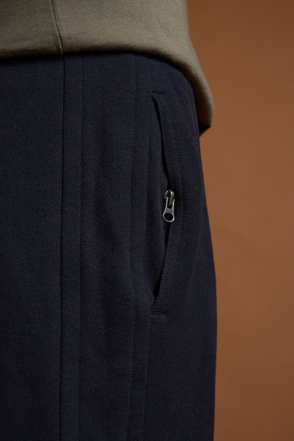 Close-up of a person wearing Barkers Heritage Track Pant in dark navy with a zippered side pocket and an olive green top, set against a brown background.