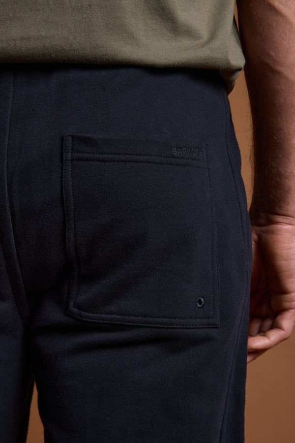 Close-up of a person in Barkers Heritage Track Pant by Barkers, dark navy with a rectangular back pocket, styled with an olive green shirt against a brown background. A relaxed hand at their side enhances the casual vibe.
