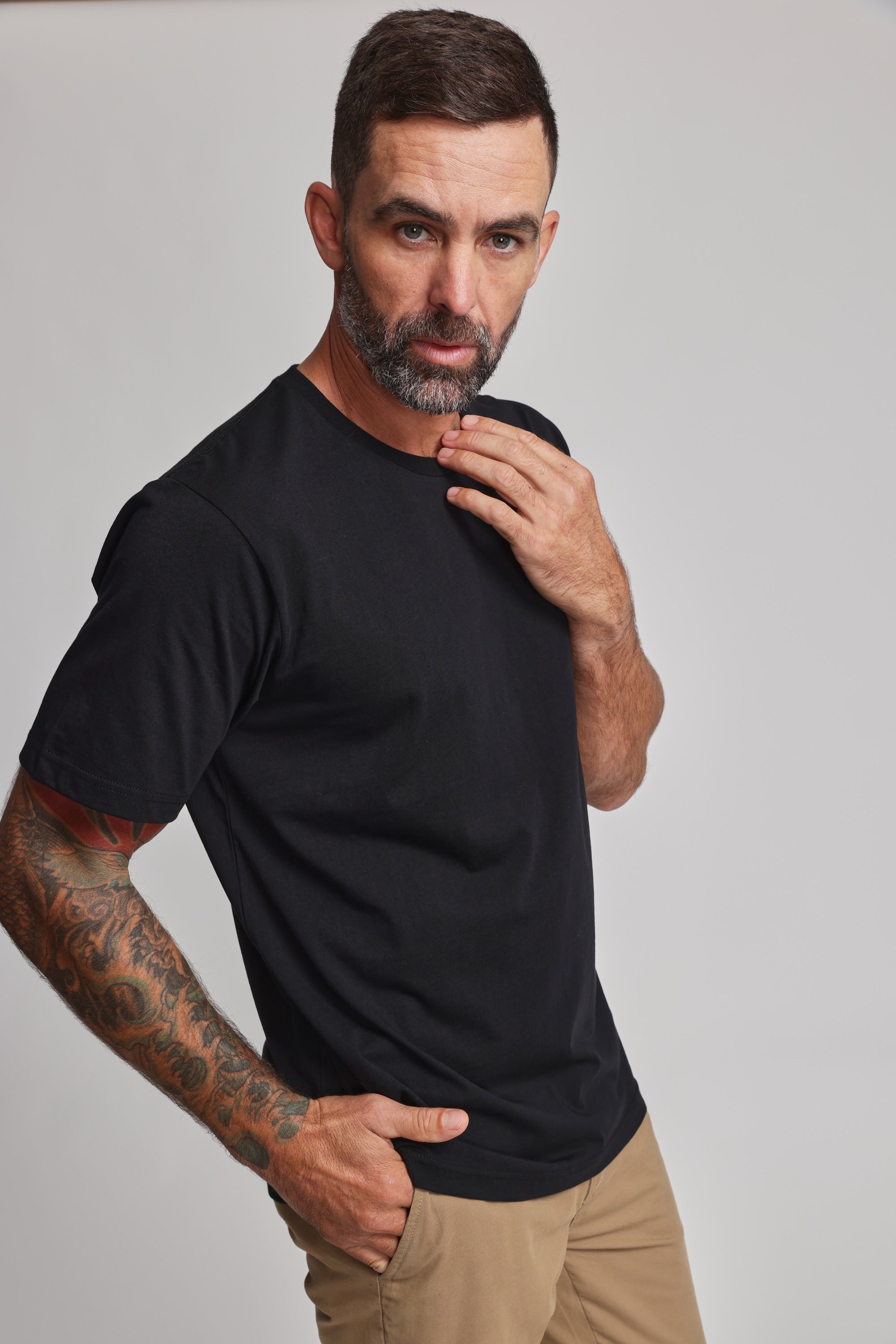 A man with short brown hair, a beard, and a tattoo sleeve poses against a plain light background wearing the Barkers Organic Crew Tee in black—a 100% organic cotton mid-weight shirt—with beige pants. One hand is on his chin, the other in his pocket.