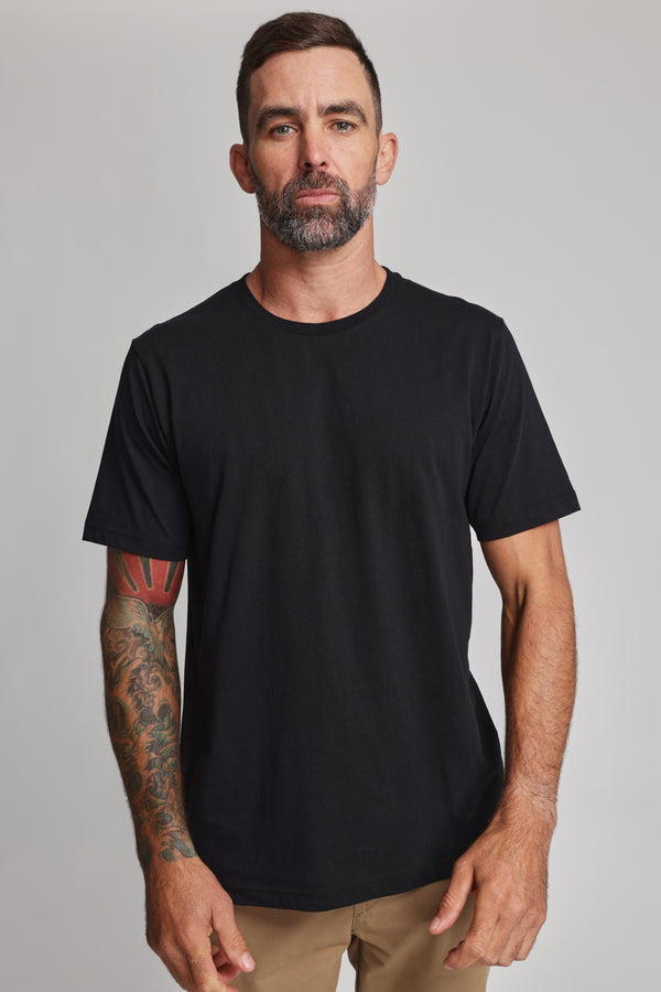 A man with short dark hair, a beard, and tattoos on his right arm wears the Barkers Organic Crew Tee—plain black and made from 100% organic cotton—with tan pants, standing against a light gray background and looking at the camera neutrally.