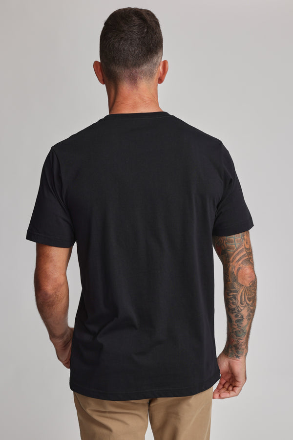 A man with short dark hair and a tattooed right arm stands with his back to the camera, wearing Barkers' Organic Crew Tee in black—crafted from 100% organic cotton—and khaki pants, set against a plain light background.