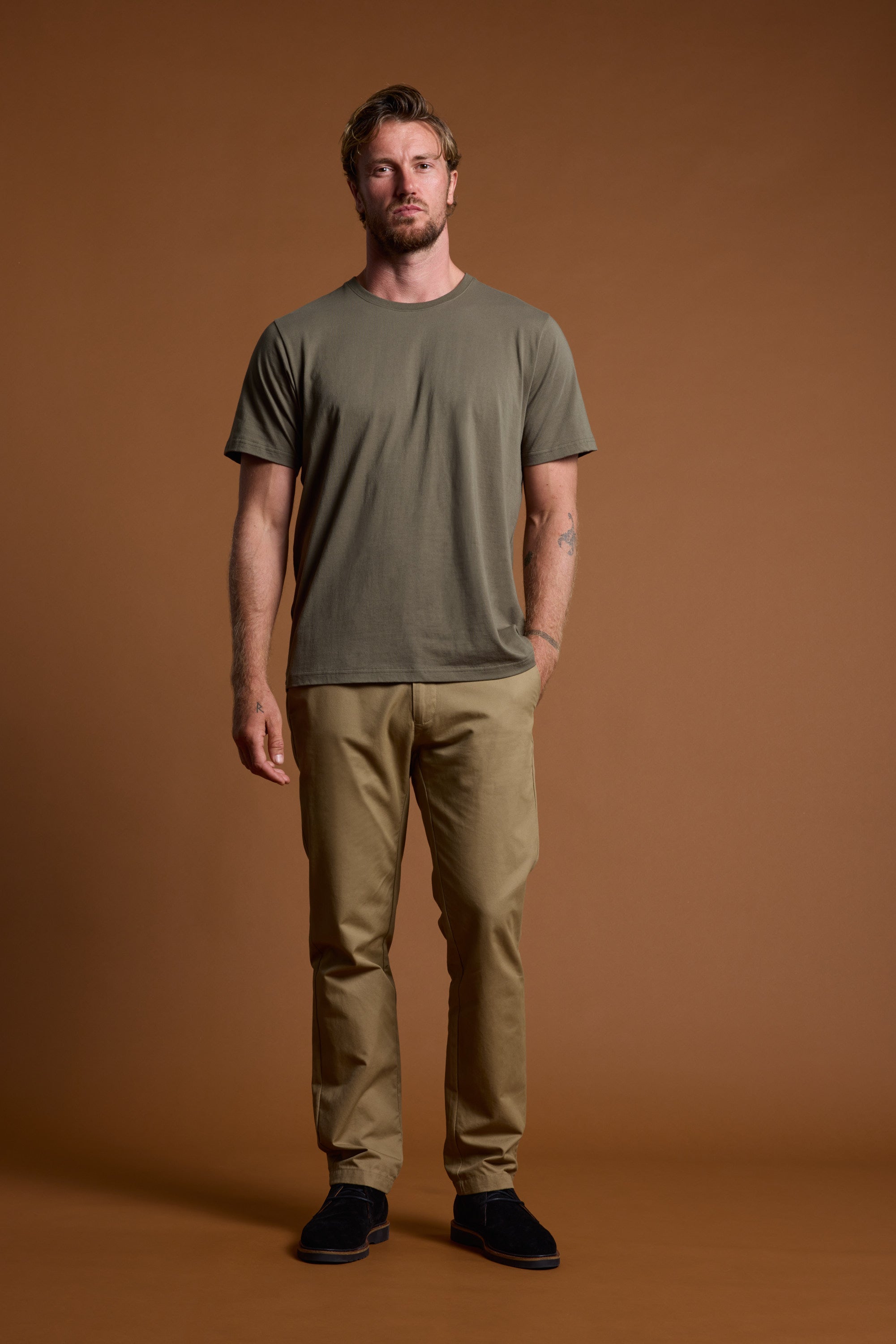 A man stands against a plain brown background, wearing the Barkers Organic Crew Tee in khaki green—crafted from 100% organic cotton—paired with tan pants and black shoes. He has short hair, a neutral expression, and his hands rest at his sides.
