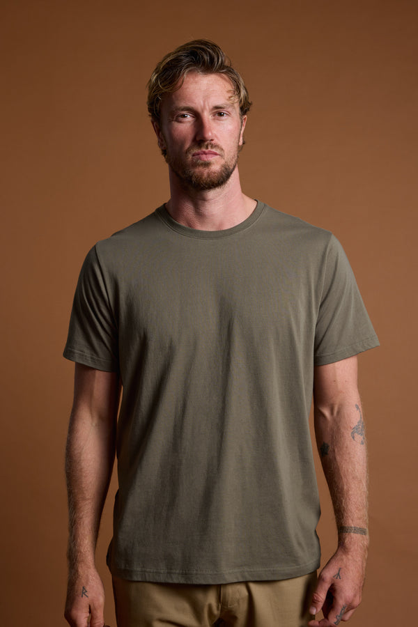 A man with short, wavy hair and a beard wears the Barkers Organic Crew Tee in olive green, made from 100% organic cotton, with beige pants. He stands against a brown background with a neutral expression and visible arm tattoos.
