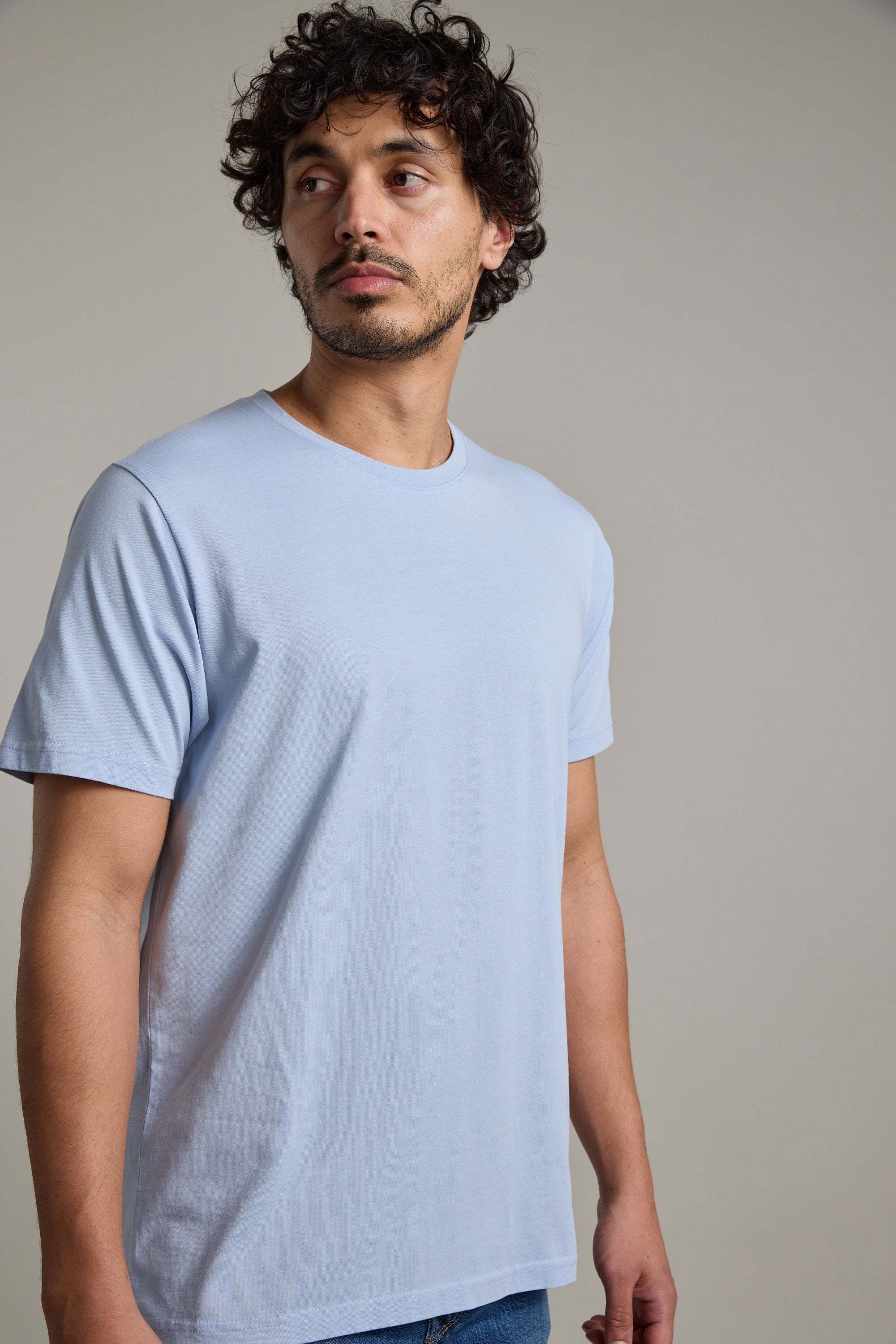 A man with curly hair and facial hair wears a Barkers Organic Crew Tee in light blue and blue jeans, standing against a plain light gray background, looking slightly to his left.