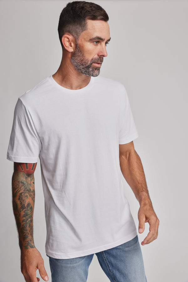 A man with short dark hair and a beard wears the Barkers Organic Crew Tee, made from 100% organic cotton, along with light blue jeans. He stands against a light gray background, displaying a colorful tattoo on his left arm.