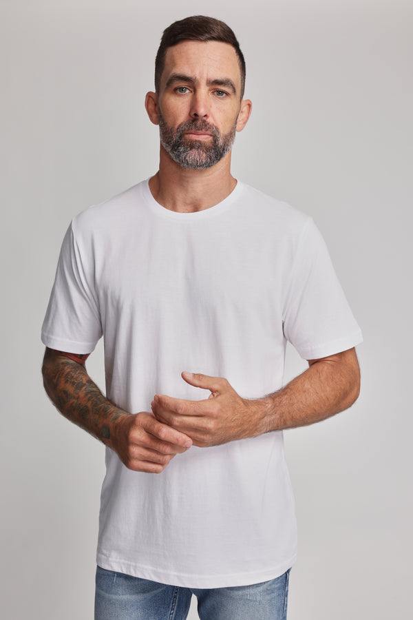 A man with short dark hair and a beard, arms tattooed, stands facing forward in the Barkers Organic Crew Tee (100% organic cotton) and light blue jeans, wearing a neutral expression against a plain light background.