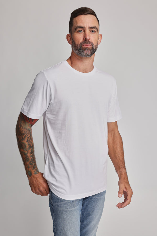 A man with a short beard and tattoos on his left arm wears the Barkers Organic Crew Tee, made from 100% organic cotton, and light blue jeans, standing against a plain light background.