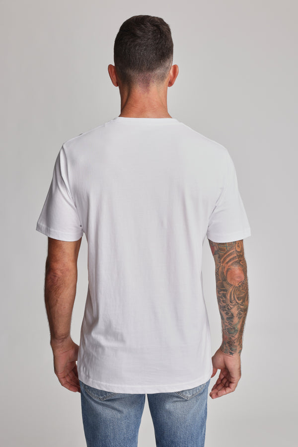 A man with short dark hair and a sleeve tattoo on his right arm wears the Barkers Organic Crew Tee in plain white (100% organic cotton) and blue jeans, standing with his back to the camera against a light, plain background.