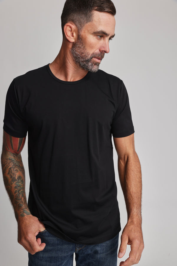 A man with short dark hair, a beard, and tattoos on his left arm wears the Barkers Linton Supima Tee in black with blue jeans. He looks down to his left against a plain light background.