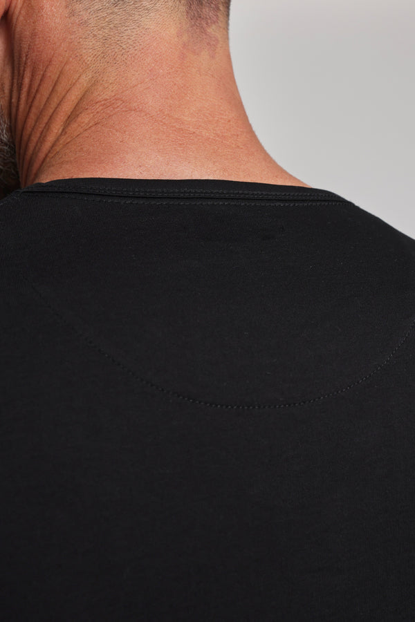 Close-up of a person’s neck and shoulders in a Barkers Linton Supima Tee, plain black crew neck style. The skin above the collar is visible against a neutral background.
