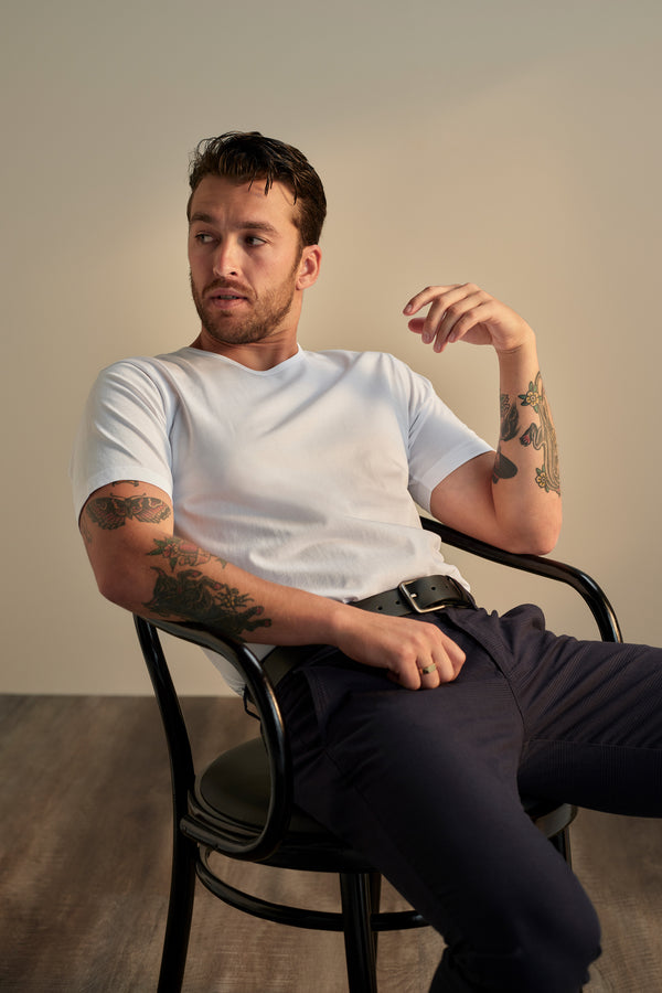 A tattooed man sits on a black chair in dark pants and the white Linton Supima Tee from Barkers. Relaxed, he looks to the side with one arm on the chair’s back and the other on his leg—a true wardrobe staple.