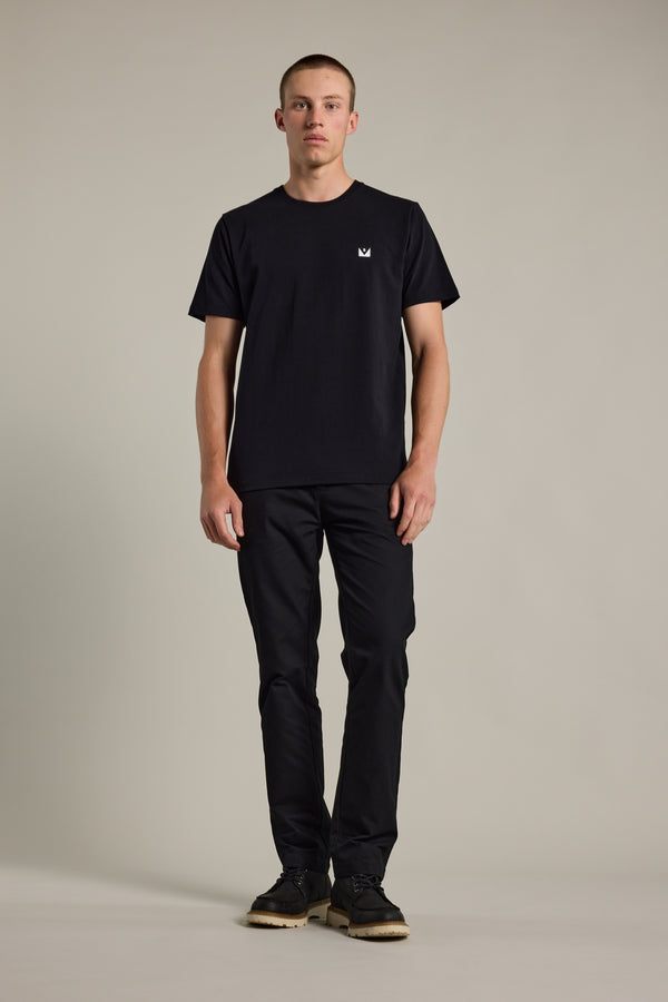 A man stands facing forward against a plain background, wearing the Barkers Newton Washed Tee—a black oversized box fit shirt with a small white logo on the chest—paired with black pants and black shoes.