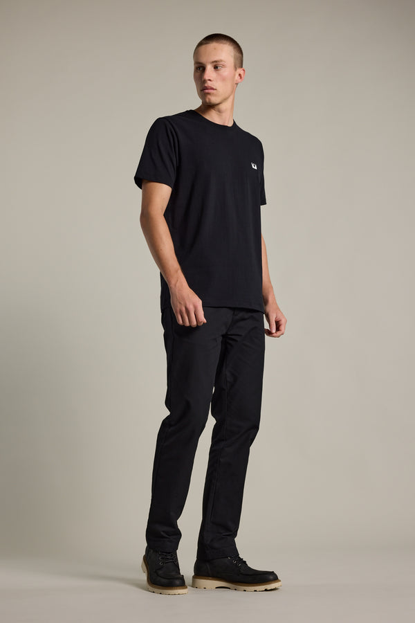 A man stands against a plain light background, wearing the Barkers Newton Washed Tee in an oversized box fit, paired with black pants and black shoes, looking off to the side with a neutral expression.