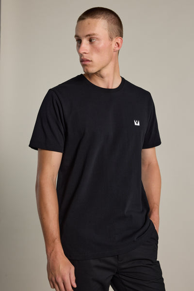 A young man wears the Barkers Newton Washed Tee, an oversized black shirt with a small white crown logo on the chest, standing against a neutral background looking left with one hand in his pocket.